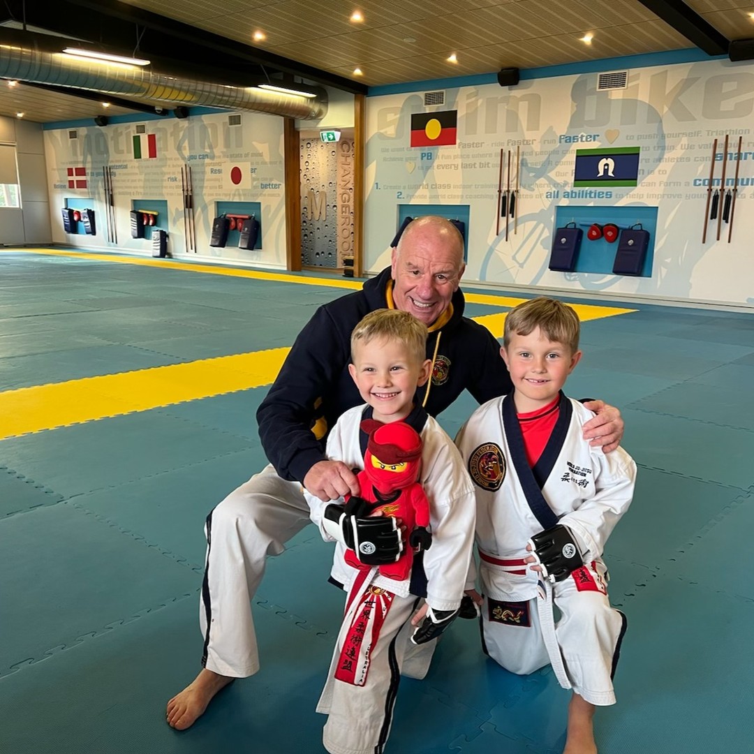 A male trainer poses with two young boys in martial arts attire in a gym with boxing bags visible in the background.