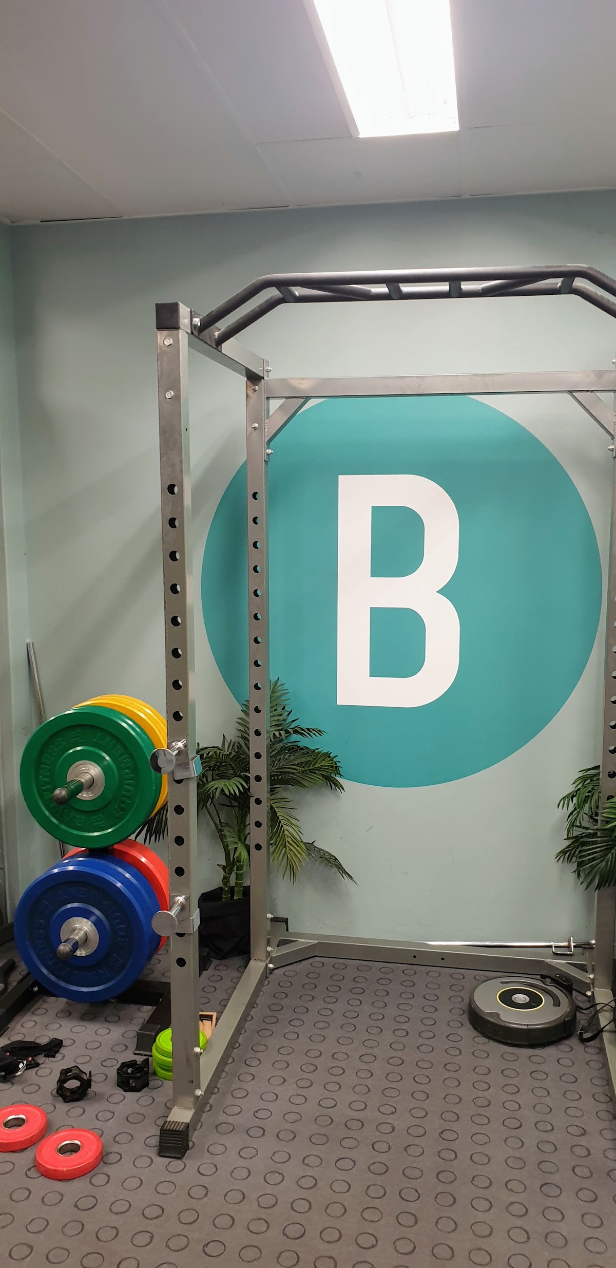 The photo shows a weightlifting area with a squat rack, weights, and a logo on the wall.