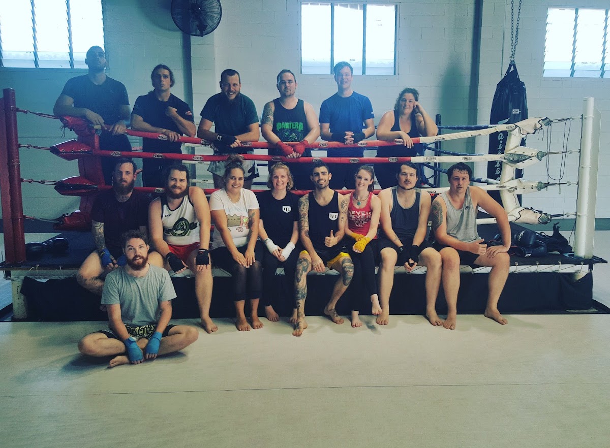 A group of mixed gender individuals are posing for a photo in front of a boxing ring and MMA cage at what appears to be a martial arts gym.