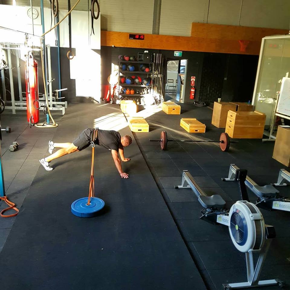 A man performs a workout exercise involving a weighted plate and rowing machine in a CrossFit gym setting.