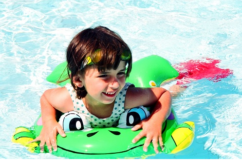 A young child is smiling and floating in a swimming pool on a green inflatable frog float.