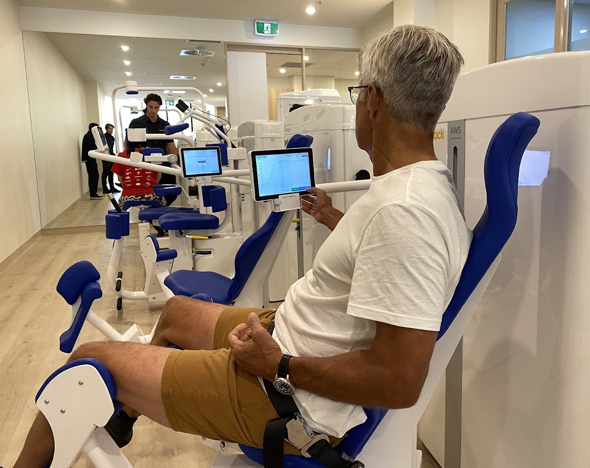 An older man is seated in a blue recovery booth with an infrared sauna, appearing to be monitoring its settings.