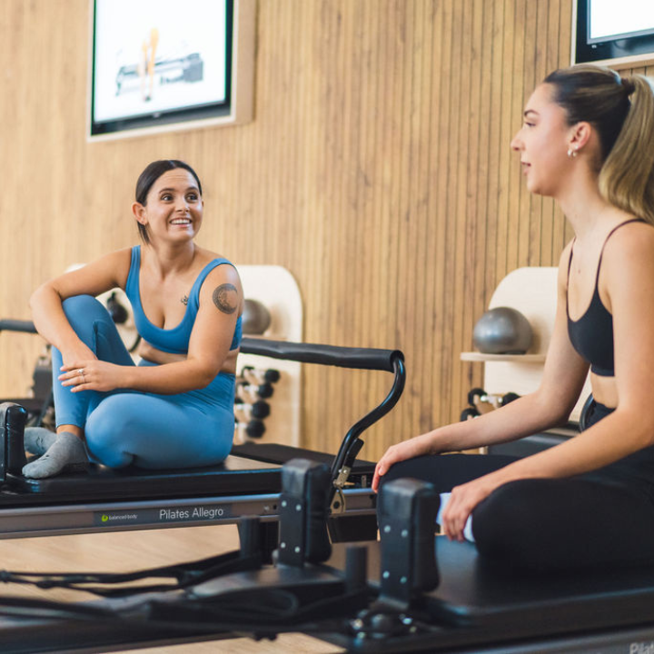 Two women are engaged in a Pilates session on reformer machines within a modern fitness studio.