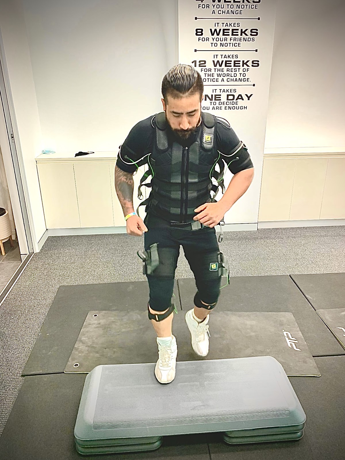 A male trainer wearing an EMS suit performs a workout on a platform in a modern fitness studio.