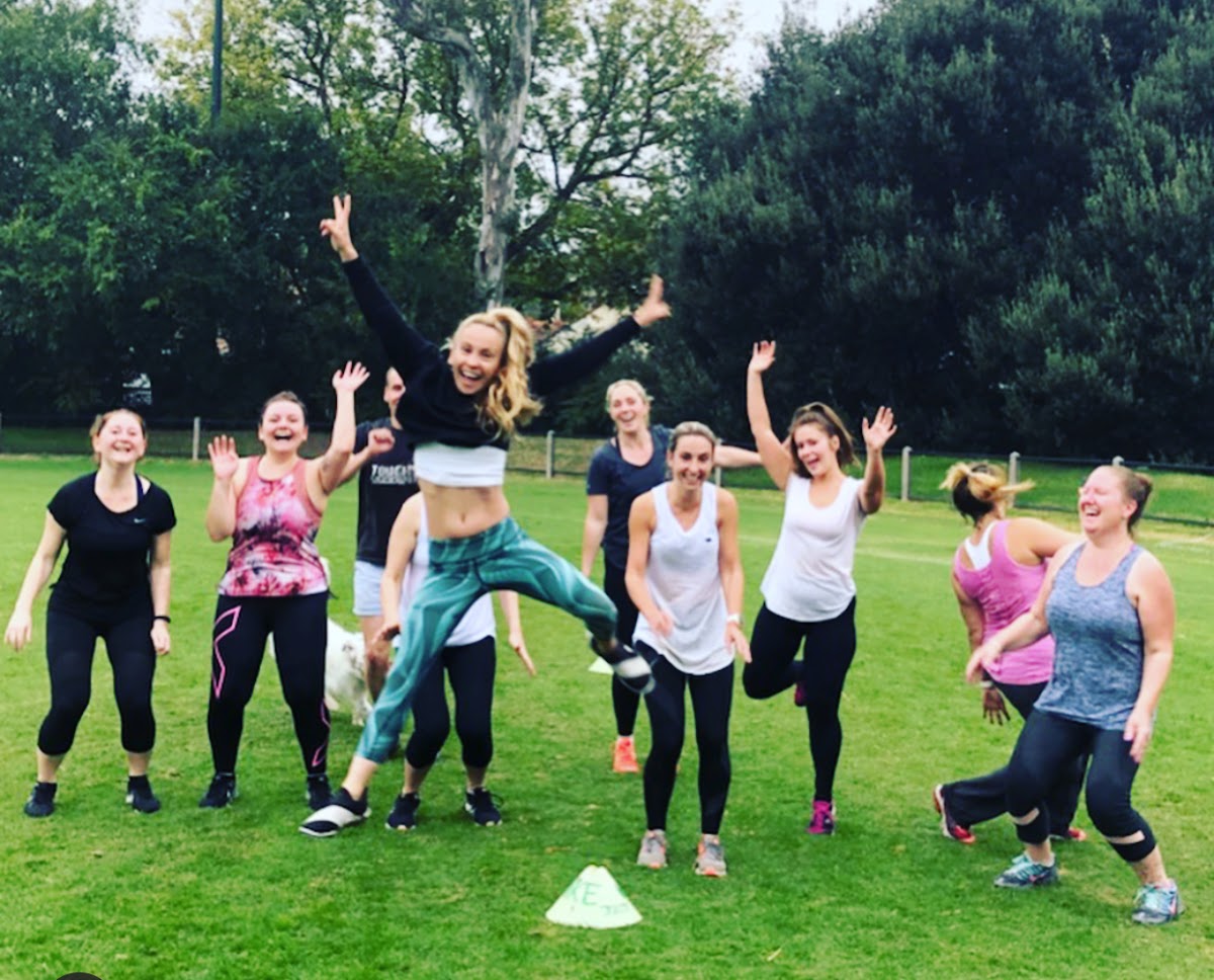 A group of women are participating in an energetic outdoor bootcamp class on a grassy field.