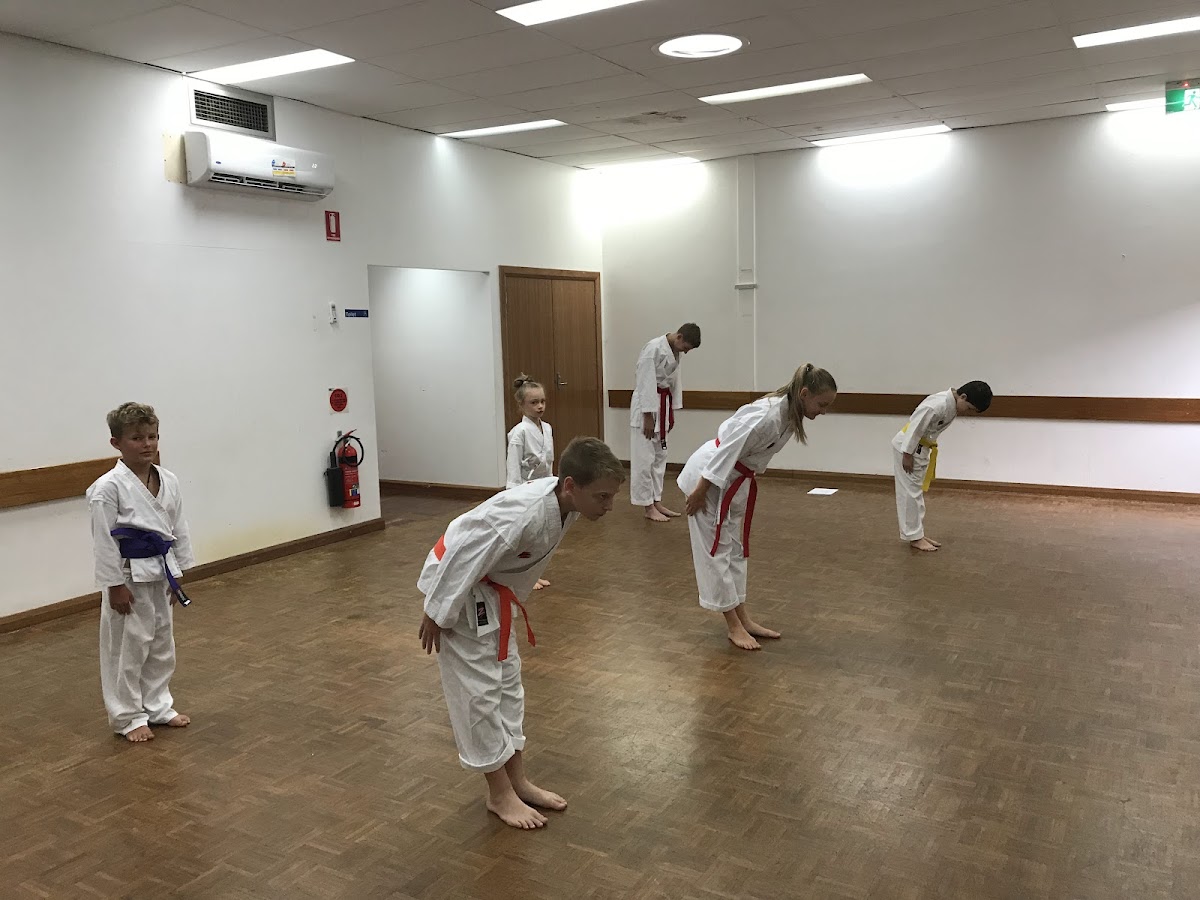 A group of children in white gis are performing a bowing exercise in a sparsely furnished martial arts studio.