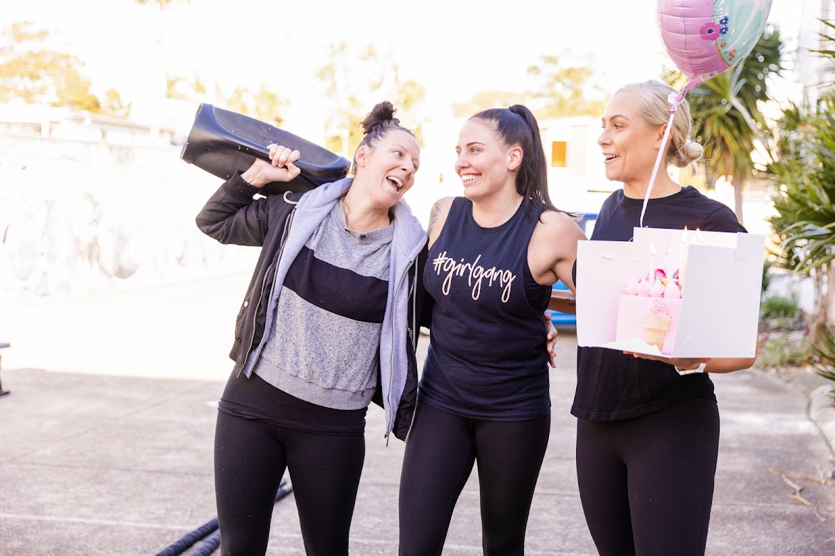 Three women, including a trainer, are laughing and posing outdoors with yoga mats, seemingly after a workout or celebration.