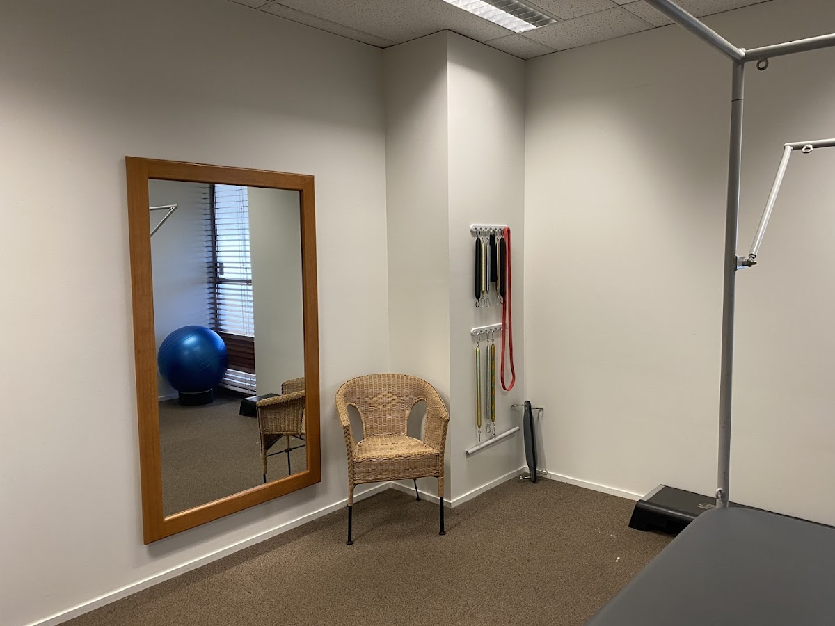 A small, minimalist studio space contains a reformer pilates machine, a mirror, a chair, and a blue exercise ball.