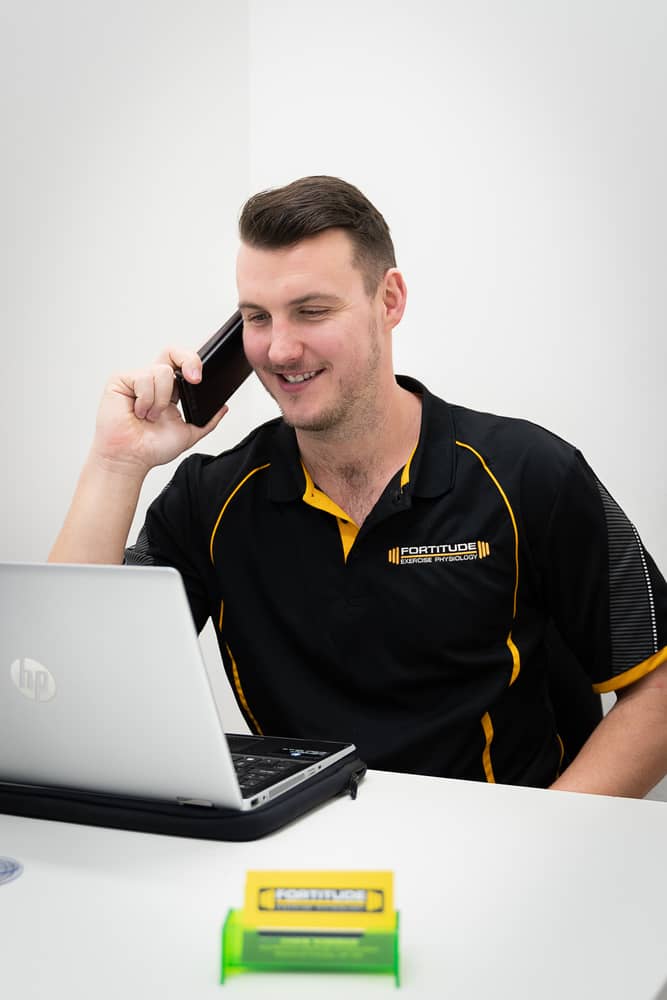 A trainer is on a phone call while working at a desk with a laptop and promotional materials.