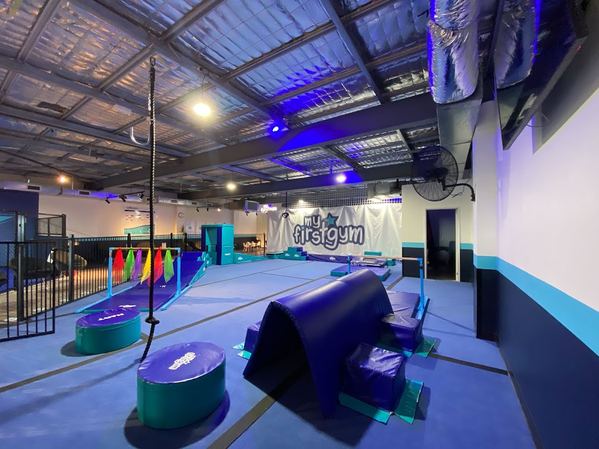 The photo shows an indoor gym space with gymnastic rings, foam blocks, and a blue and green color scheme, featuring a logo on the wall.