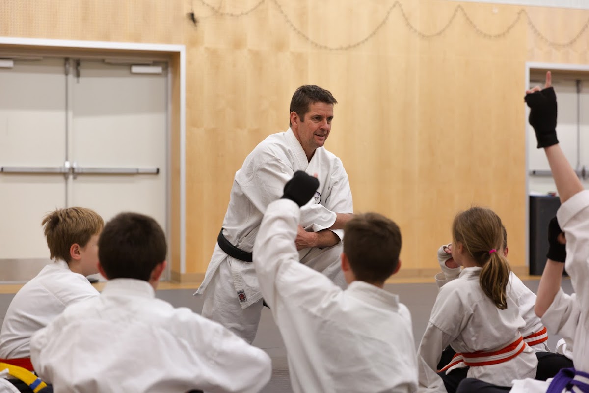 A male trainer in a white gi is instructing a group of children practicing martial arts on a mat.