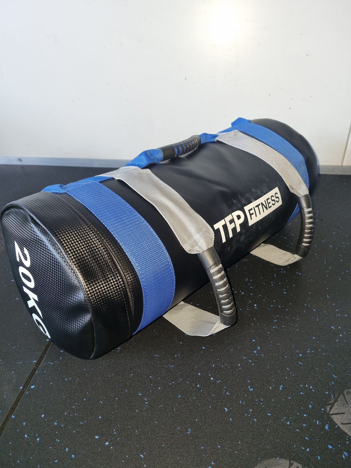 A close-up shot of a black and blue sandbag with the logo 'TFP Fitness' resting on a rubber floor.