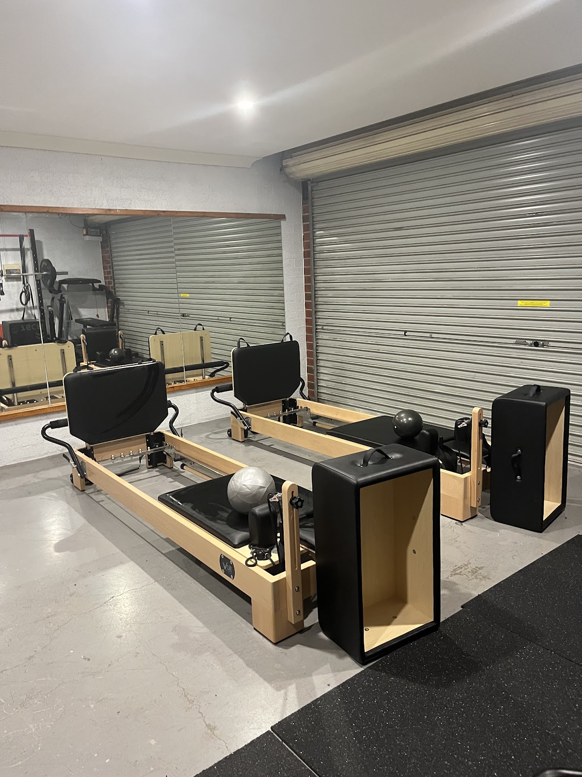 The photo shows a dimly lit gym space with several reformer pilates machines and yoga mats set up in what appears to be a garage or industrial setting