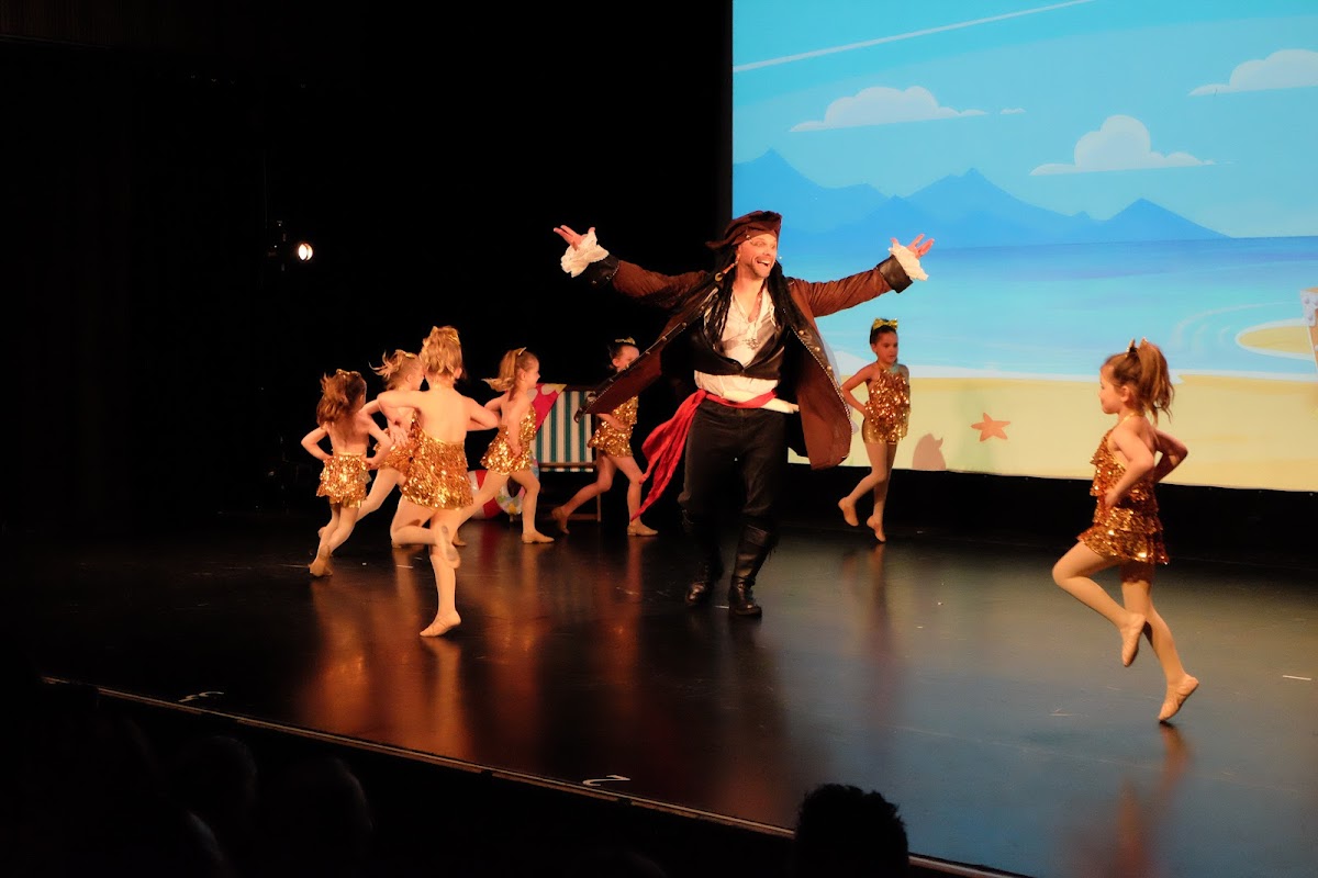 A group of young children and a female instructor are performing on a stage with a projected backdrop.
