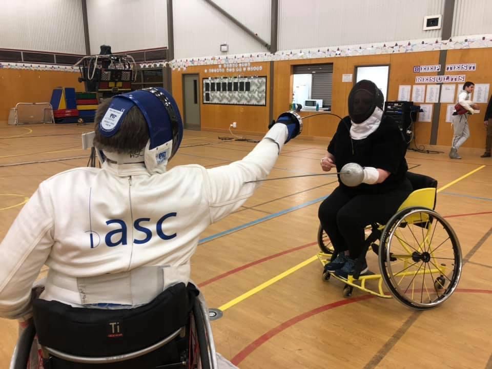 Two individuals, one in a wheelchair and wearing fencing gear, are engaged in a fencing match within a large, industrial-style gymnasium.