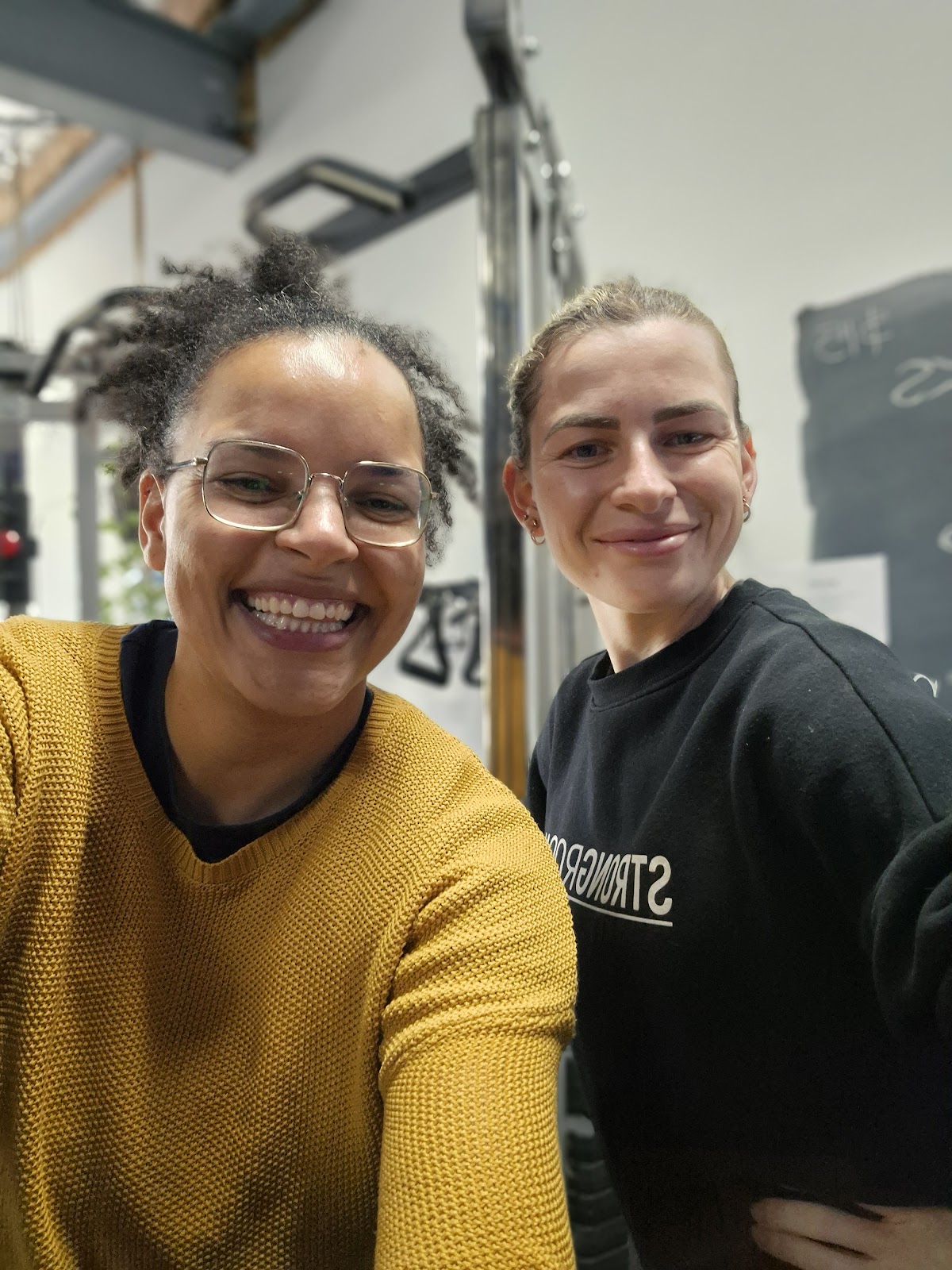 Two smiling women, one wearing glasses, pose for a photo in what appears to be a gym setting with cable machines visible in the background.