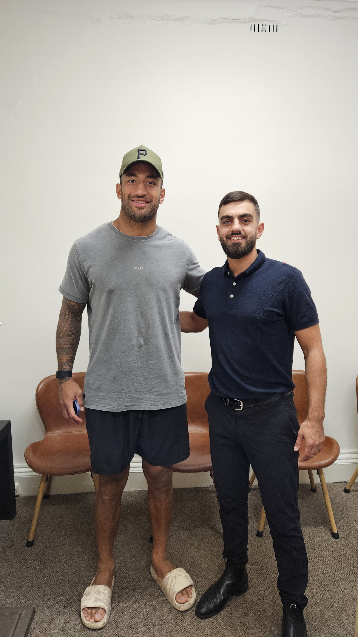 Two men, one with tattoos, pose for a photo in a sparsely furnished room with a couch.