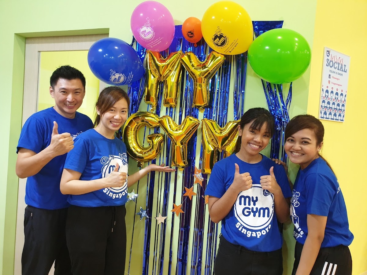 A group of four smiling individuals in matching shirts pose for a photo with balloons and decorations, seemingly celebrating an event at a gym.