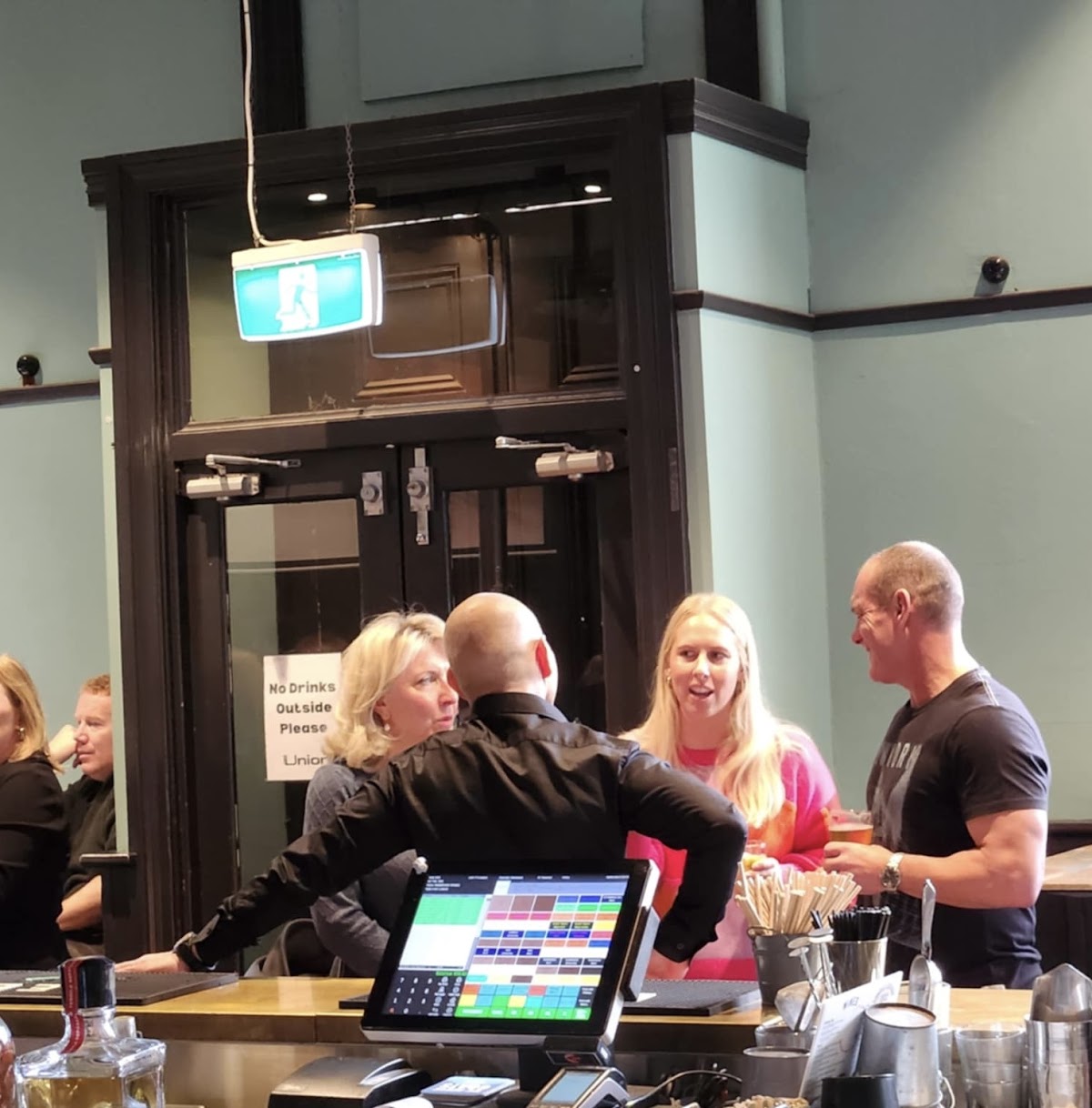 The photo shows a gym interior with people gathered around a bar area, seemingly socializing or ordering drinks.