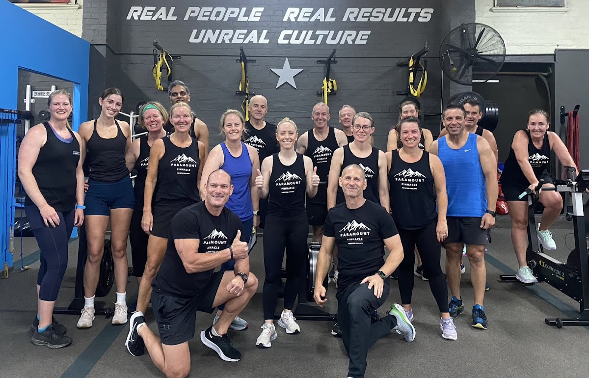 A large group of people wearing matching athletic wear pose for a photo in a gym setting with industrial elements and weightlifting equipment visible.