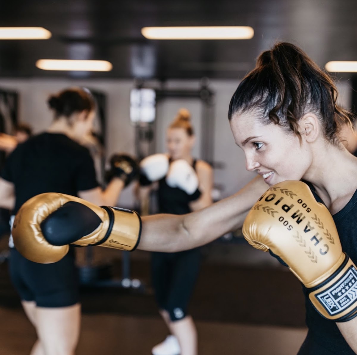 A group fitness class is taking place in a gym setting with participants practicing boxing techniques around punching bags.
