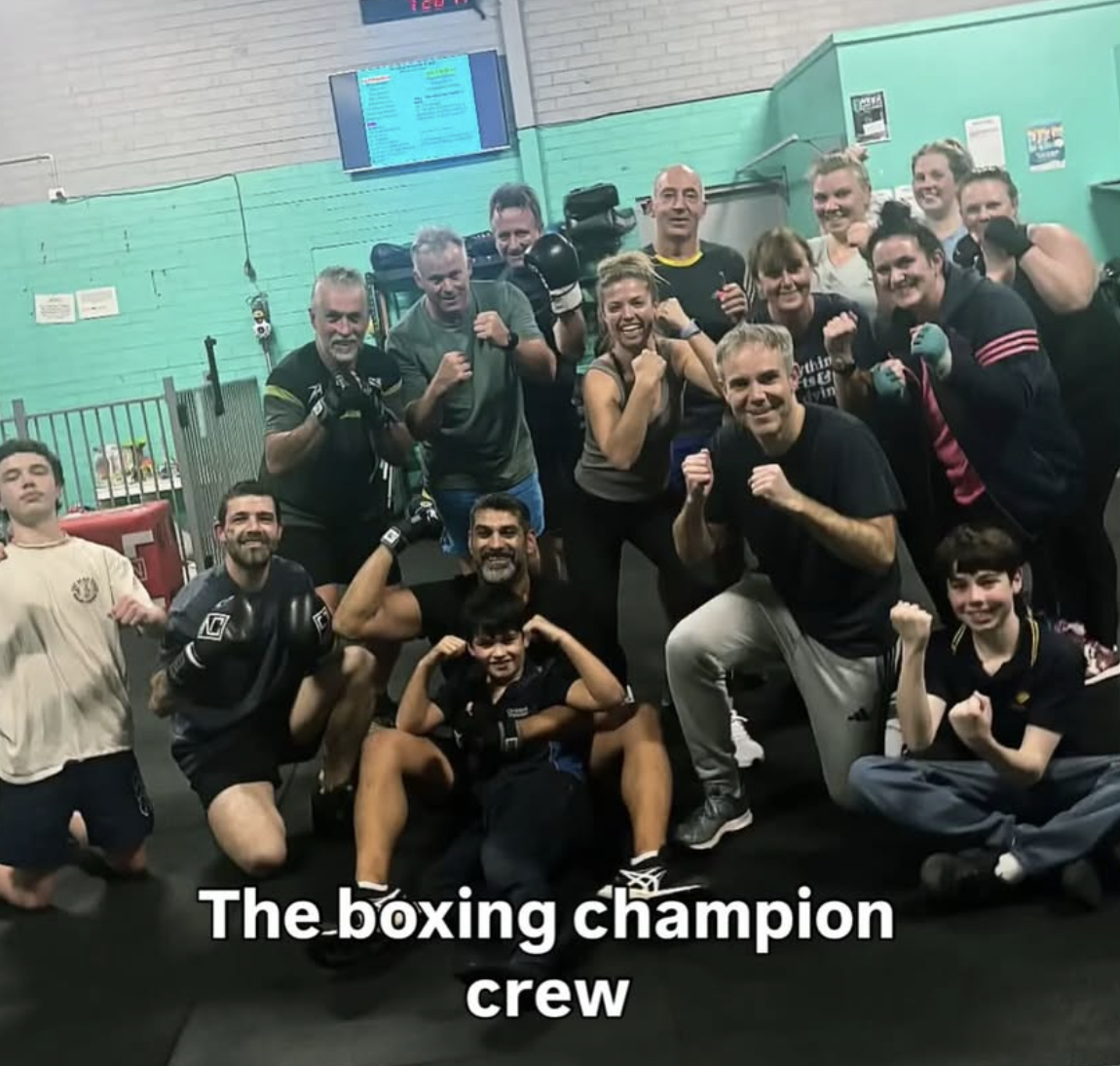 A large group of people, including a trainer, are posing for a photo in a boxing gym after a training session.