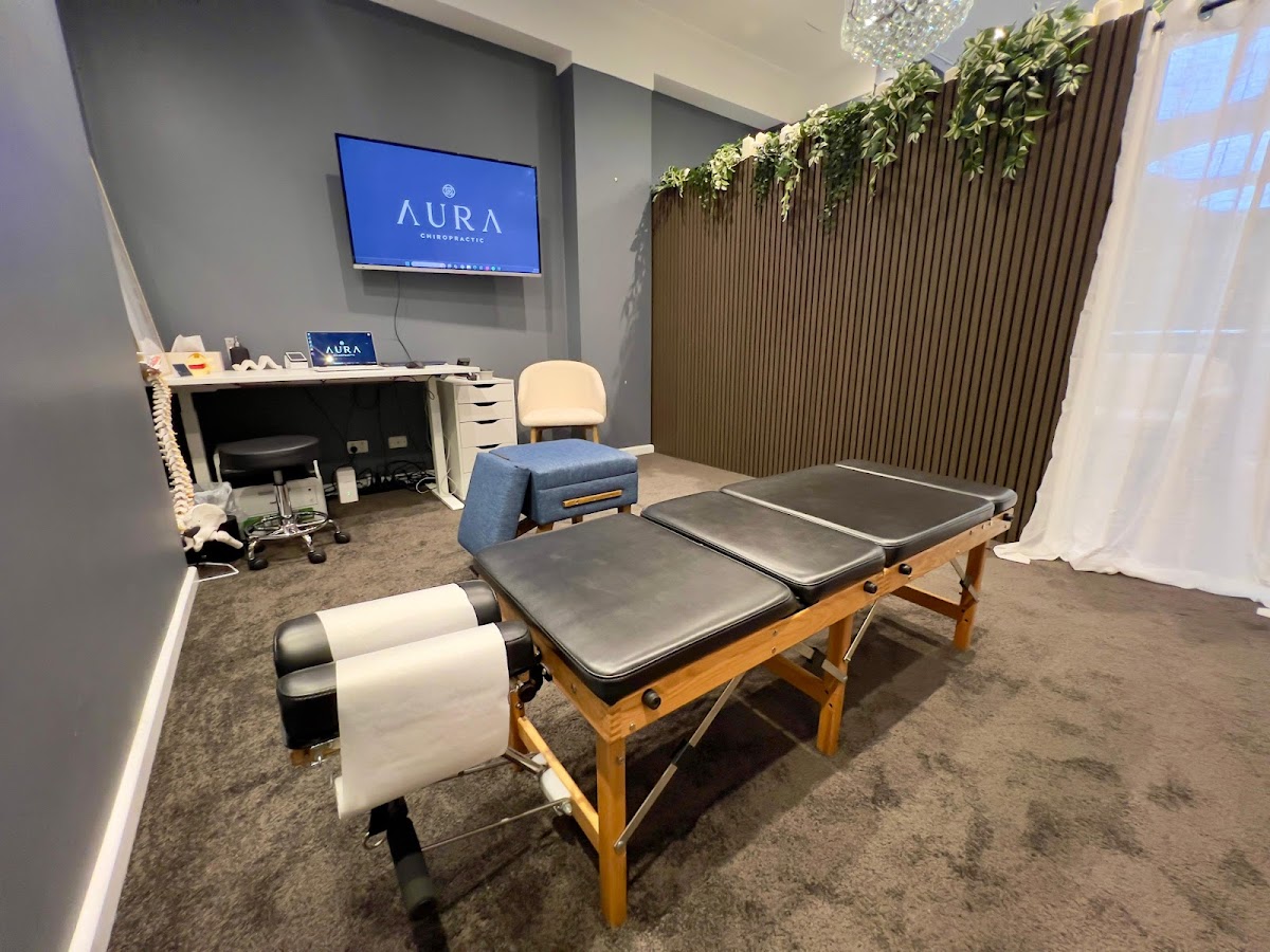 The photo shows a chiropractic studio with examination tables and equipment, featuring the Aura Chiropractic logo.