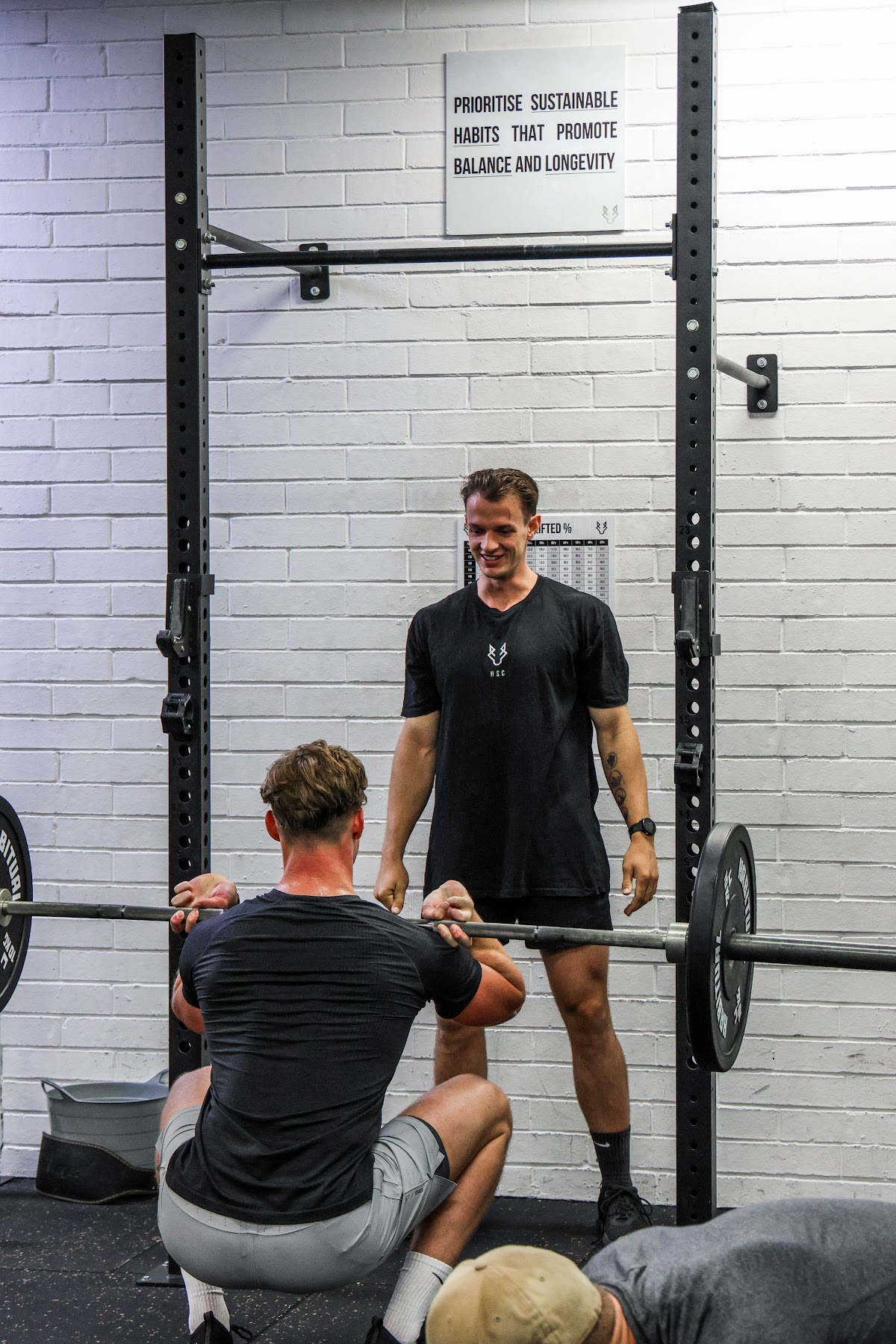 A trainer is spotting a client performing squats in a gym with exposed brick and a squat rack.