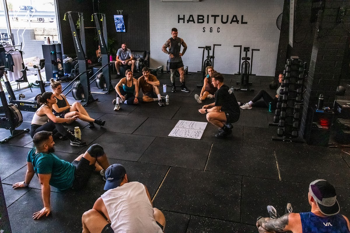 A group fitness class is taking place in a gym with rowing machines and a whiteboard displaying exercises.