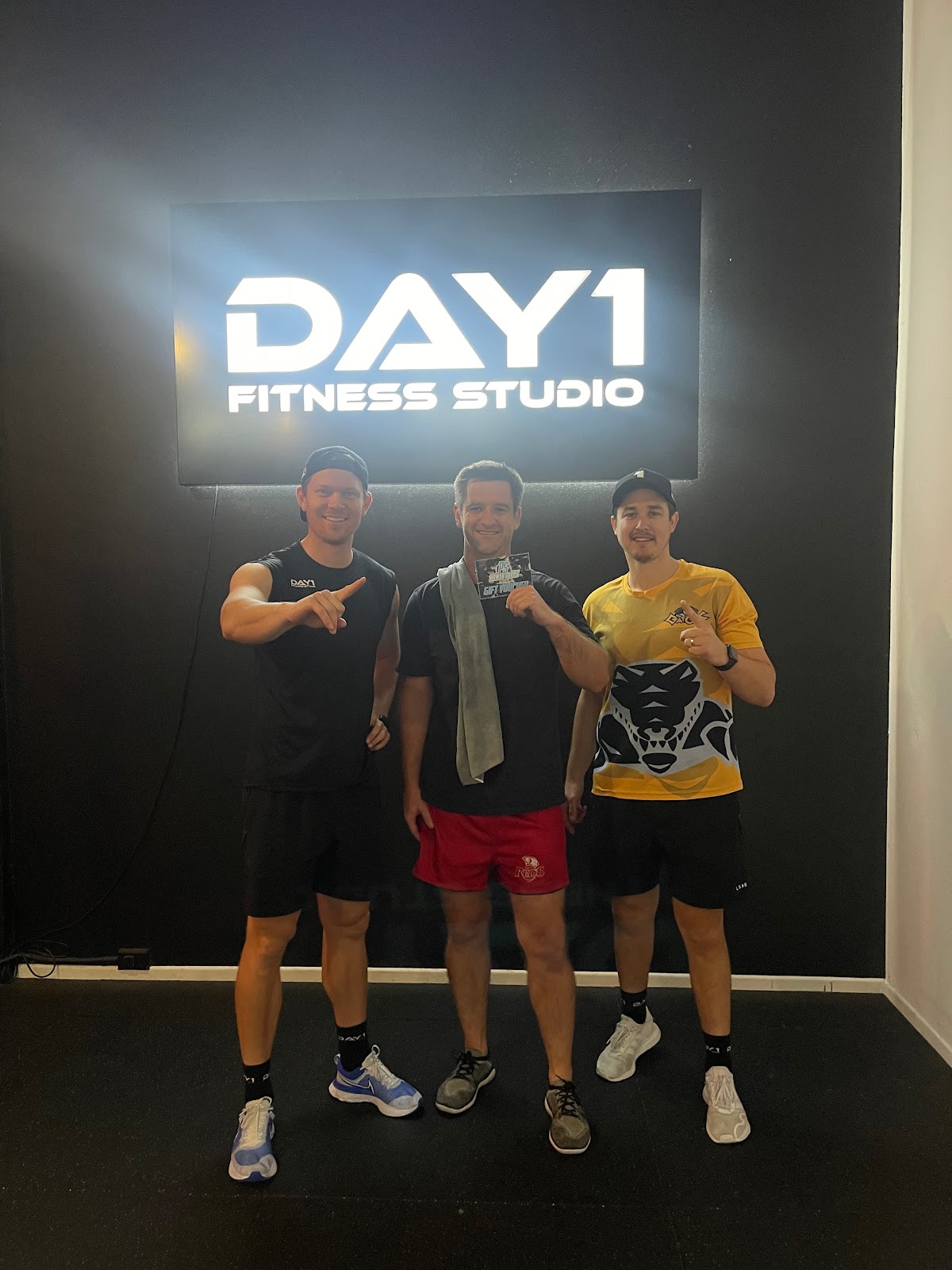 Three men pose in front of a backdrop with the 'DAY1 FITNESS STUDIO' logo, likely representing trainers or staff members.