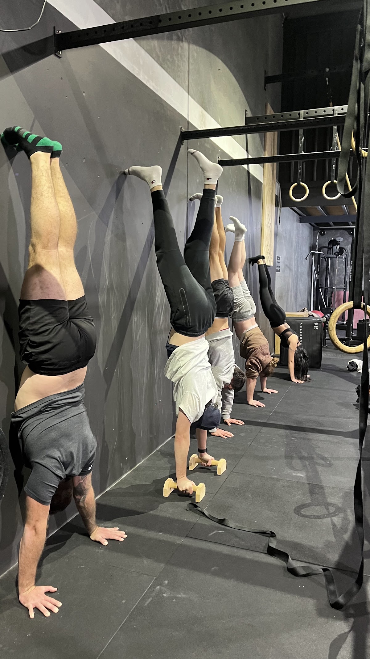 A group of people are performing handstand push-ups against a wall in a dimly lit, industrial-style gym.