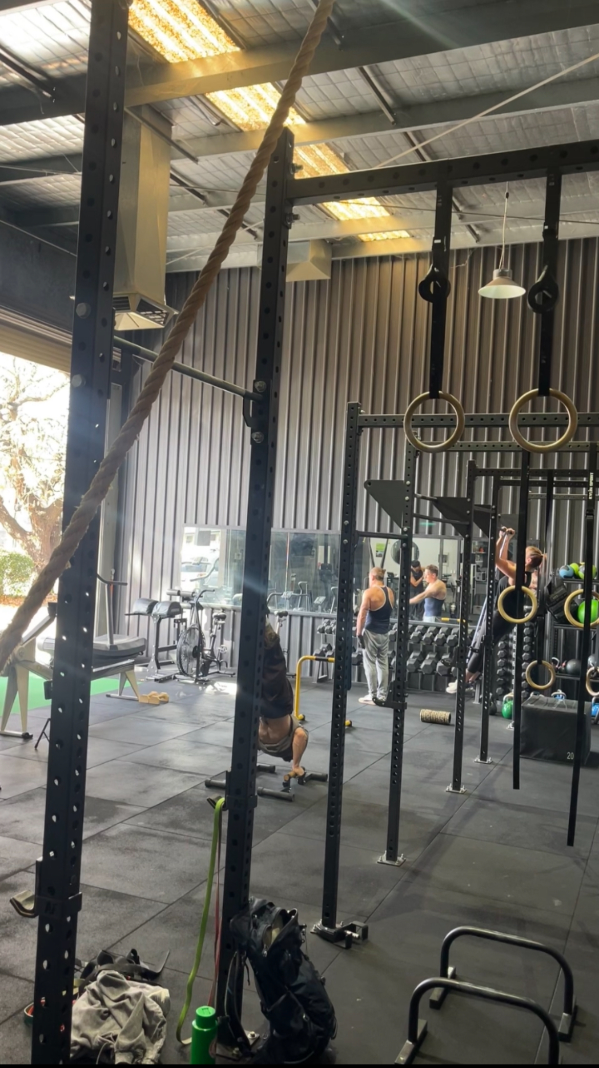 The photo shows a gym interior with a focus on a pull-up bar and gymnastic rings setup, featuring a raw industrial aesthetic and a group of people wor