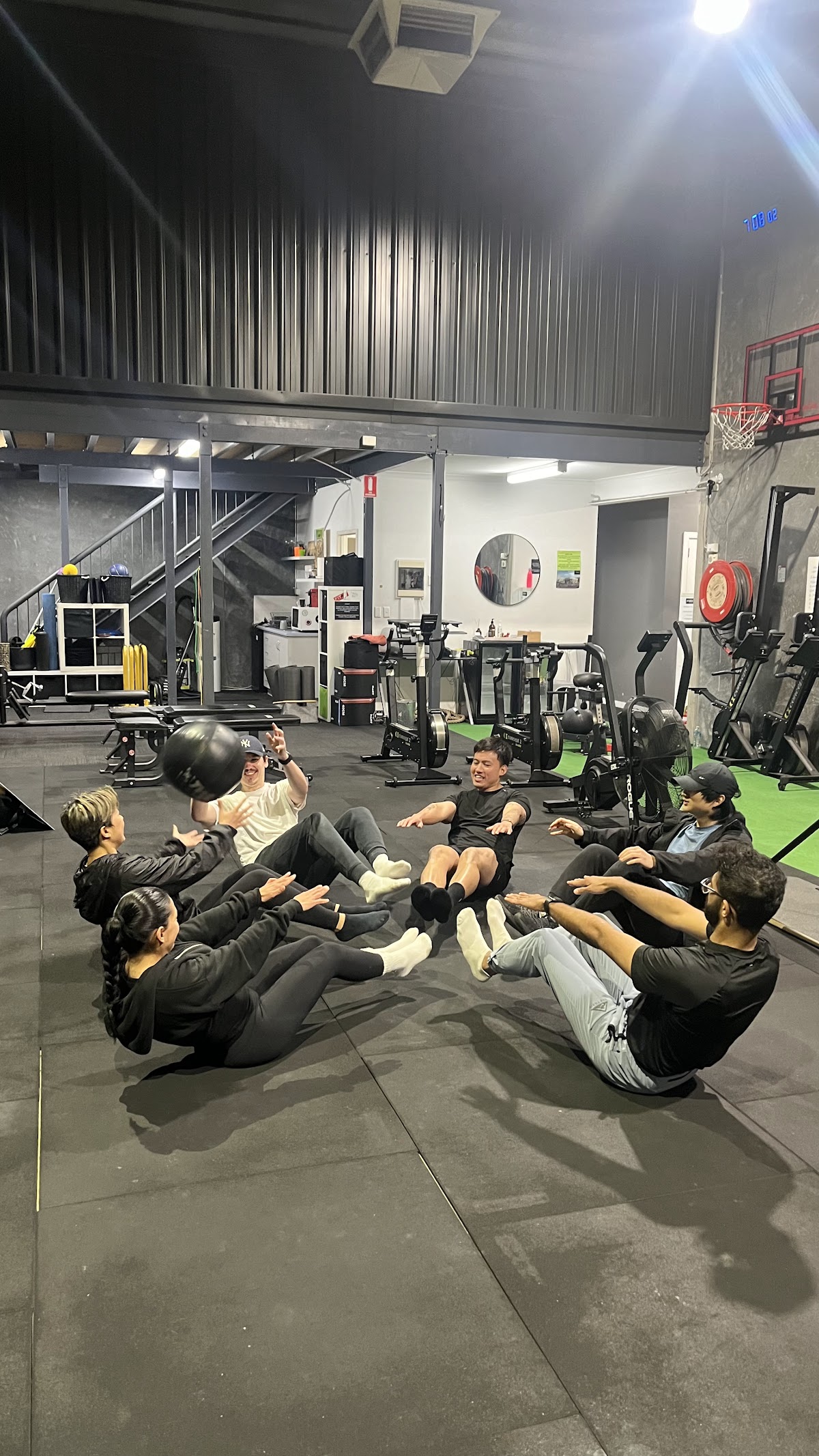 A group of people are performing core exercises with a medicine ball in an industrial-style gym.