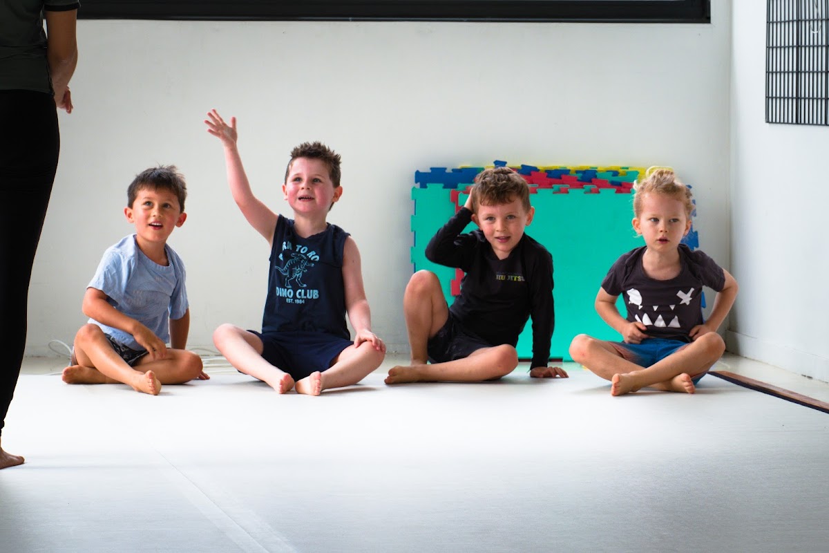 A male trainer leads a group of young children in a seated position on a yoga mat in a bright, minimalist studio.