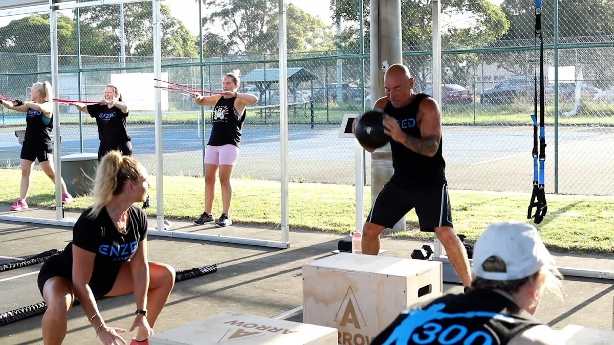 A group of people are participating in an outdoor HIIT circuit training class led by a male and female trainer.