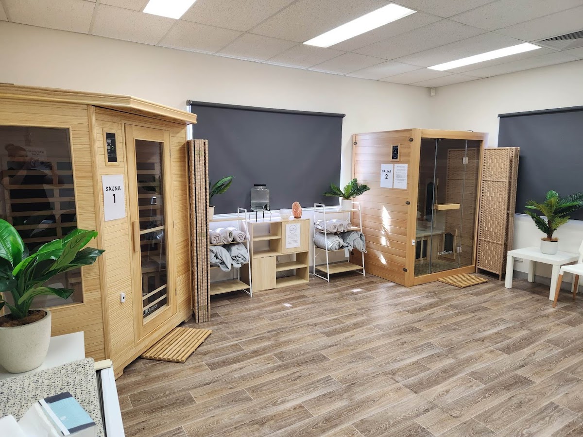 The photo shows a clean and serene interior space with two infrared saunas and a relaxation area with towels and plants.