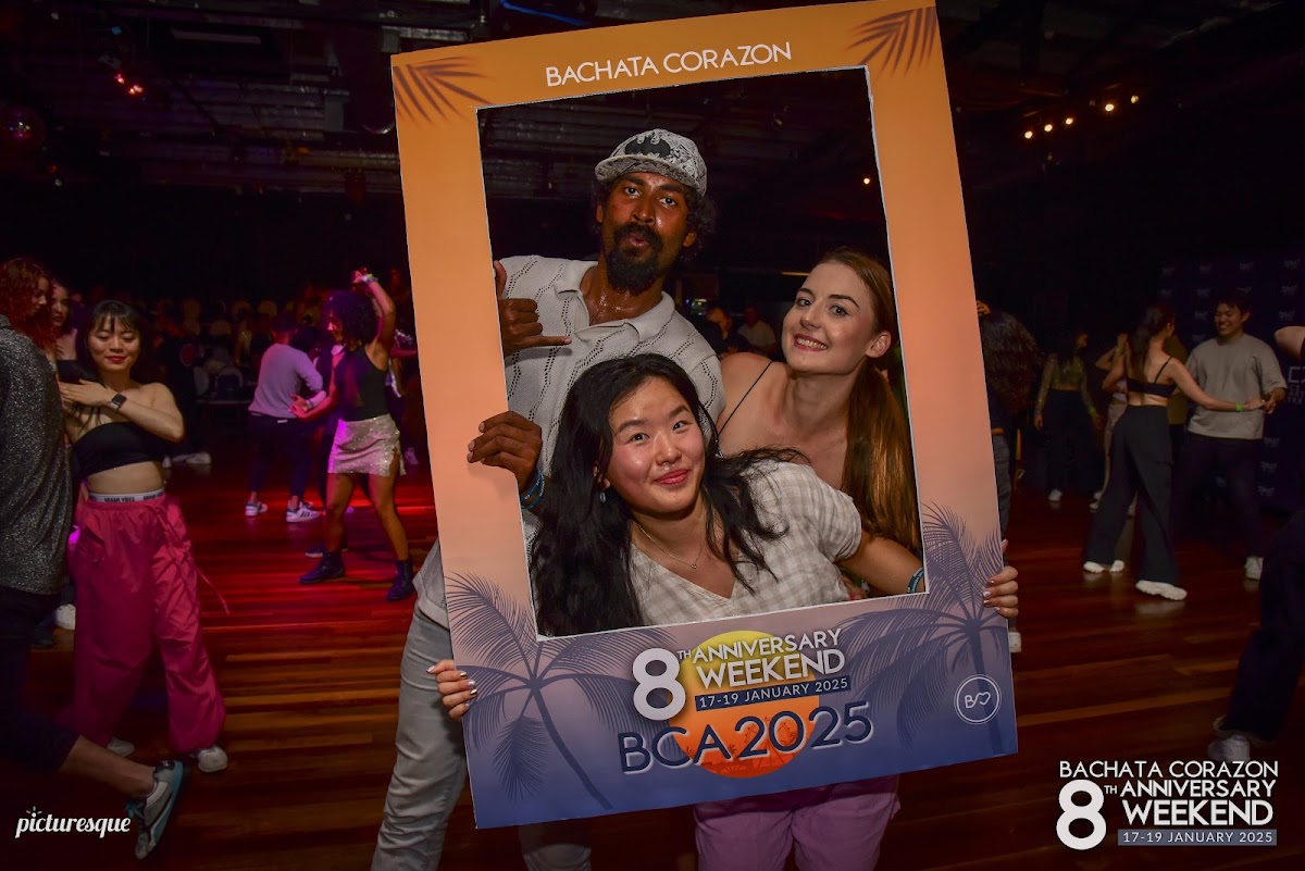 A group of people are posing for a photo at a dance event with a large frame featuring the 'Bachata Corazon' logo and anniversary details.