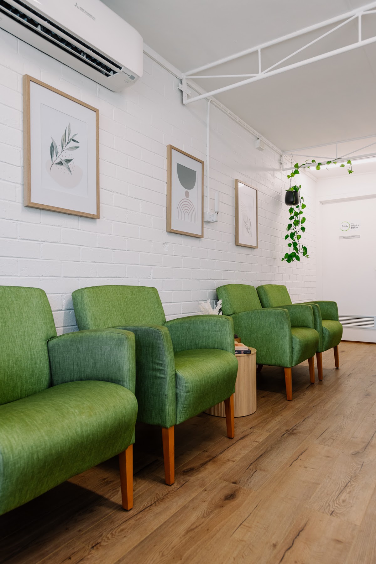 The photo shows a clean and minimalist waiting area with green chairs lined up against a white brick wall adorned with botanical prints.