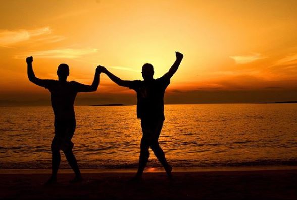 Two individuals are silhouetted against a vibrant sunset on a beach, appearing to be celebrating or performing an exercise movement.