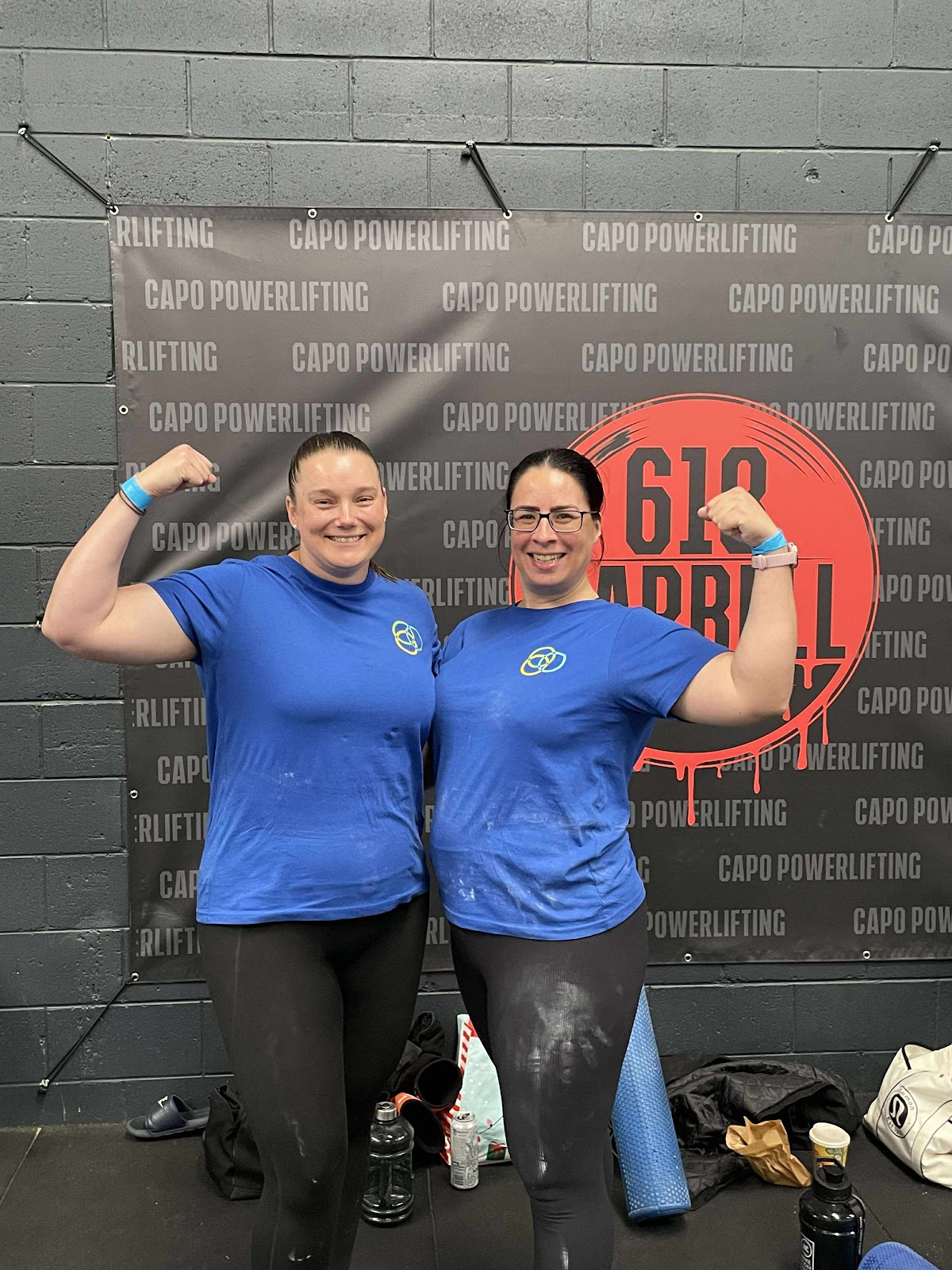 Two female trainers pose in a raw, industrial-style gym with powerlifting equipment visible in the background.