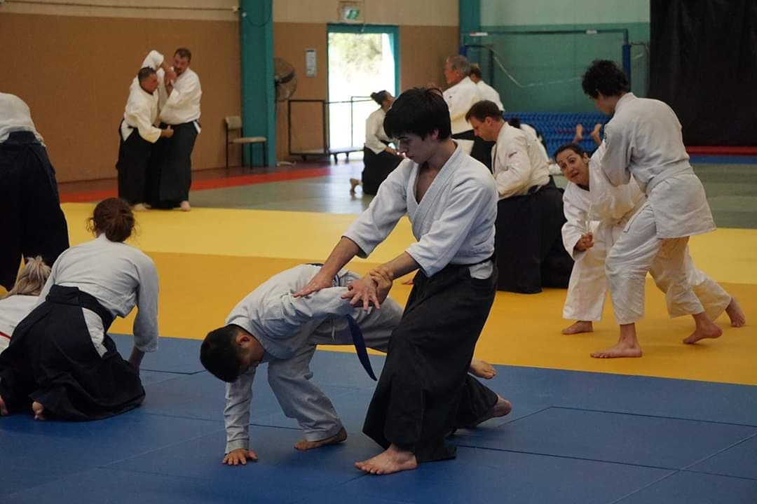 A group of people are practicing martial arts on mats in a large gym space.