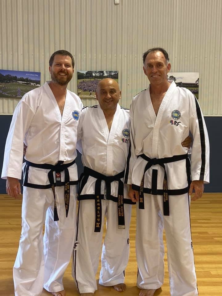 Three individuals in martial arts uniforms stand posing on a polished floor with a scenic view visible through a window behind them.