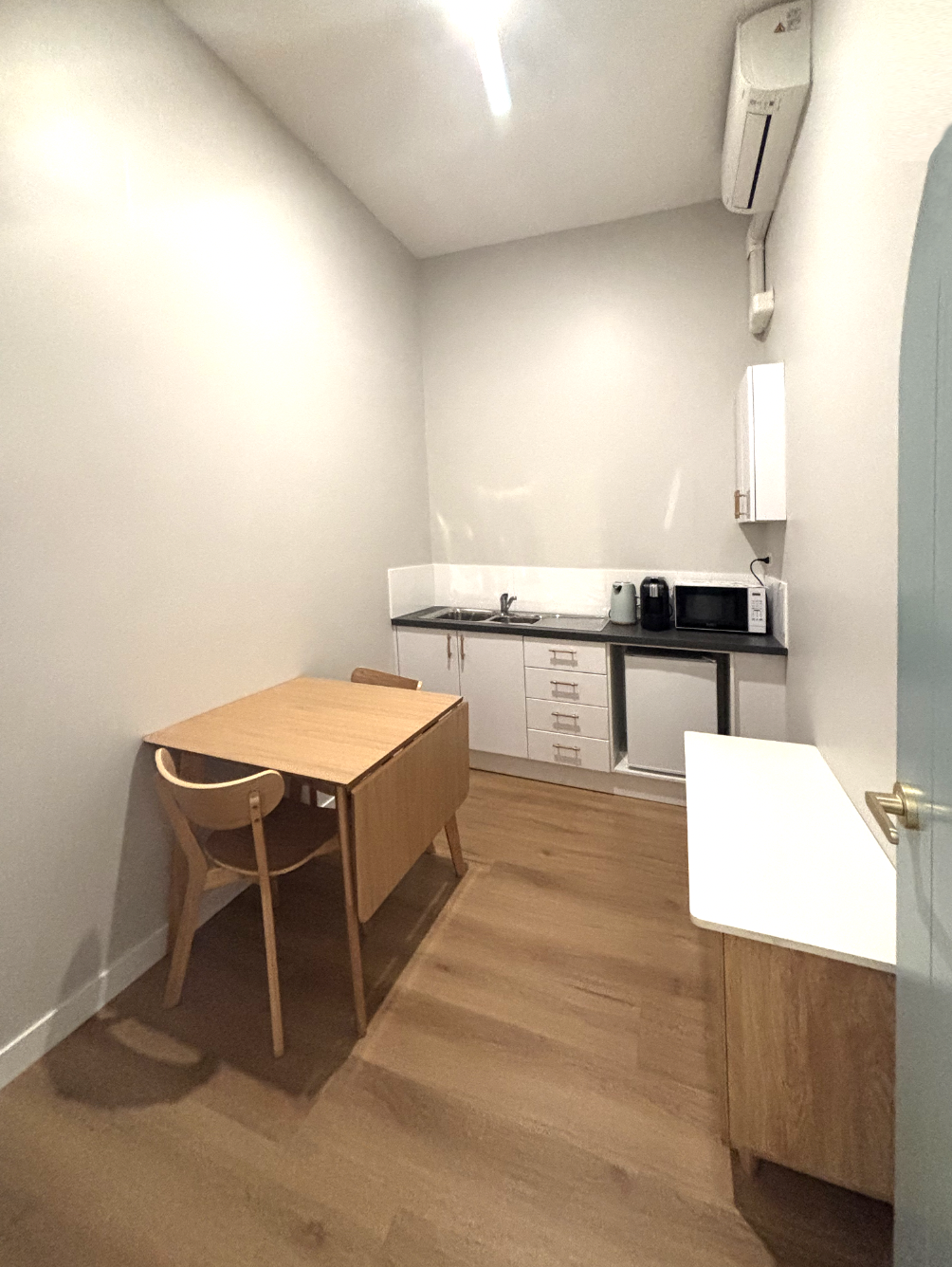 The photo shows a small, minimalist studio space with a kitchenette and a table with a chair.