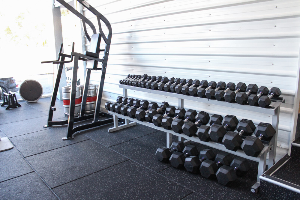 The photo shows a gym floor with rows of dumbbells on racks and a squat rack in the background.