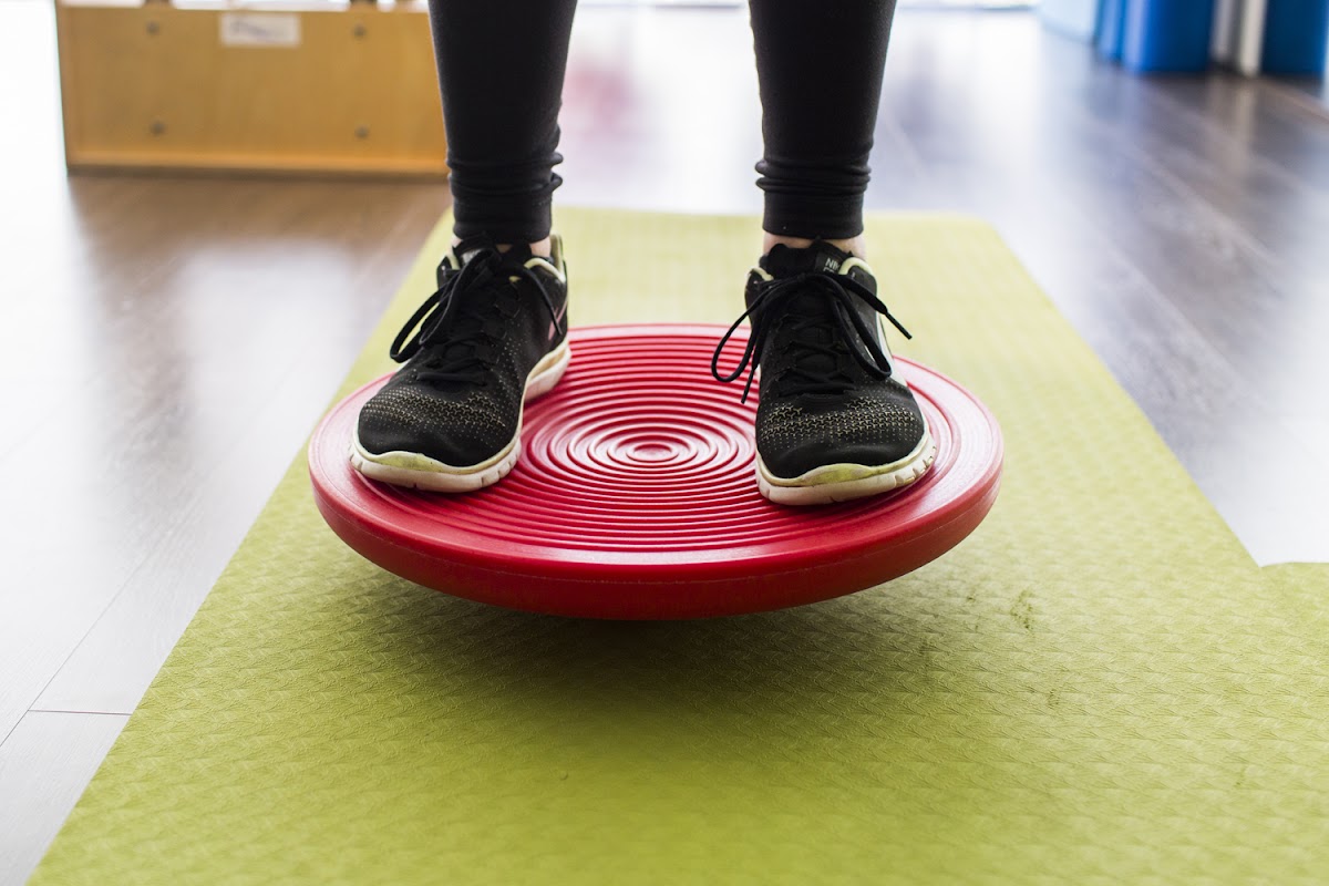 A person stands on a red balance disc atop a green yoga mat in what appears to be a fitness studio.
