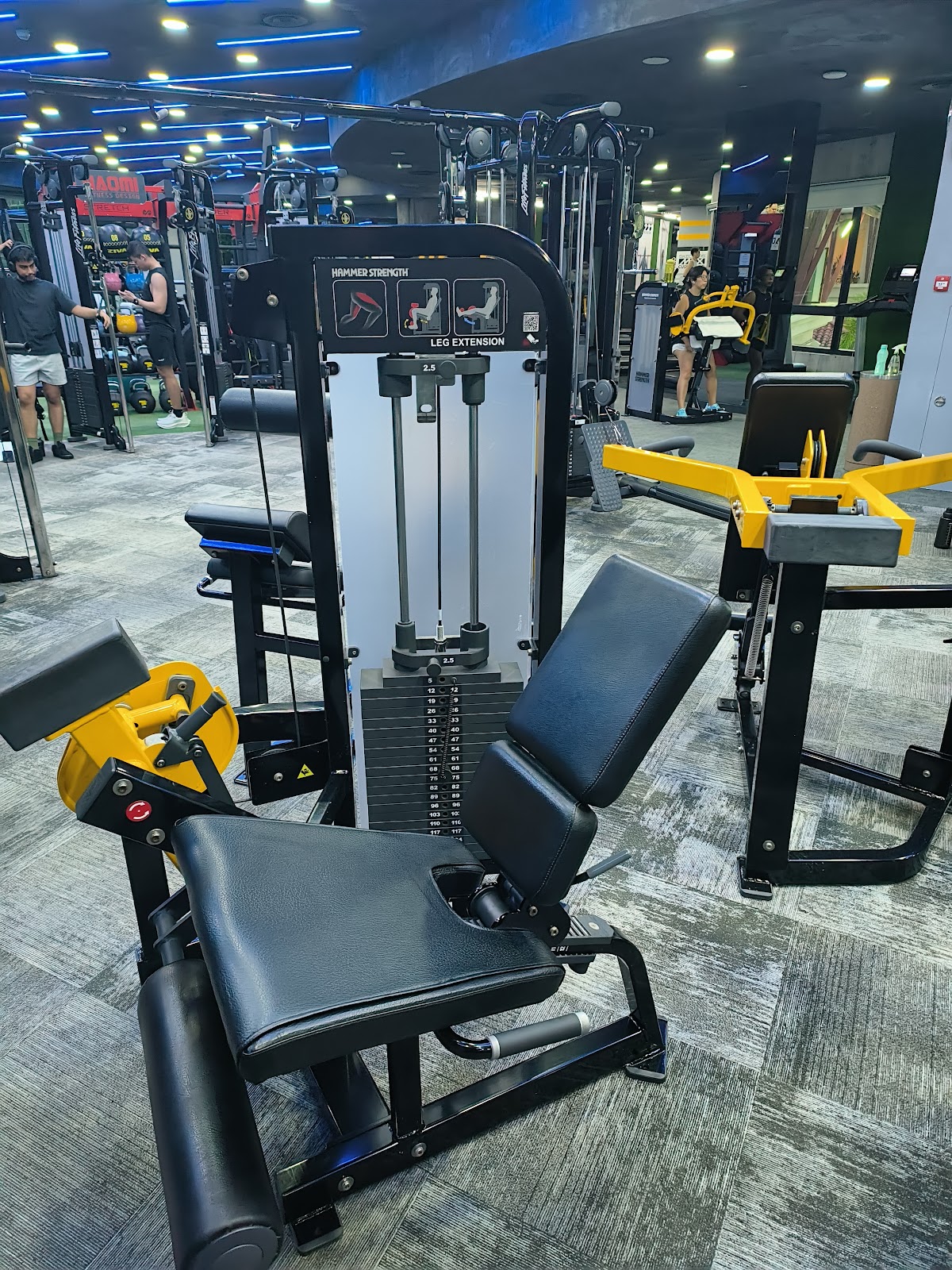 The photo showcases a well-lit gym floor with a leg extension machine in focus and other equipment visible in the background.
