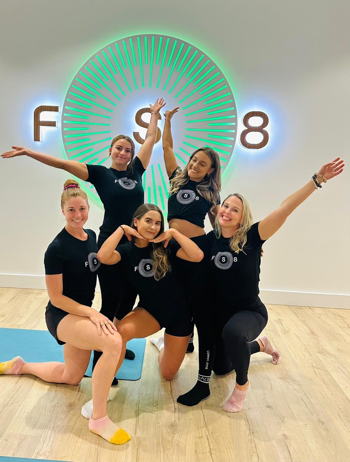 A group of women in matching workout gear pose for a photo in a brightly lit studio with a neon sign.