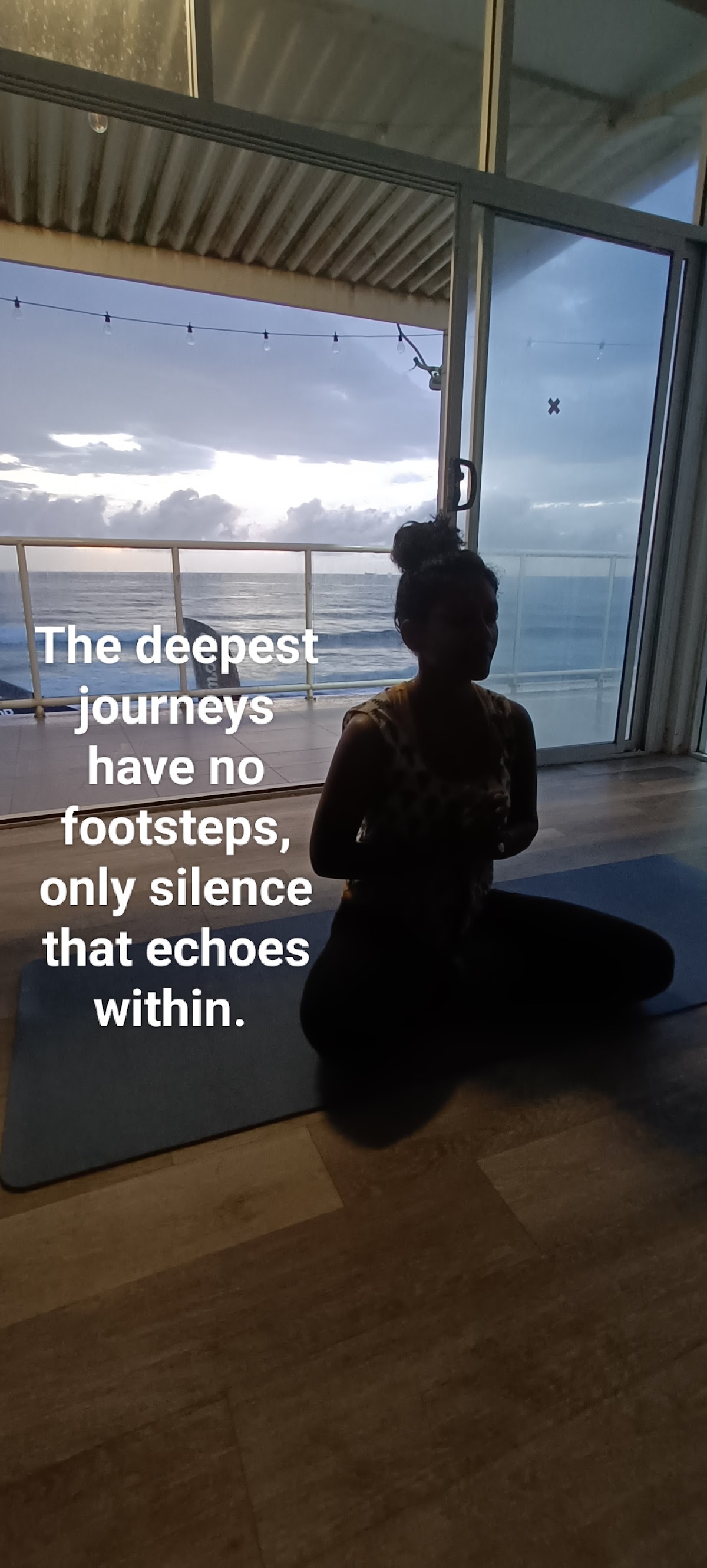 A woman in a yoga pose sits on a yoga mat in a studio with large windows overlooking a cloudy sky and ocean.