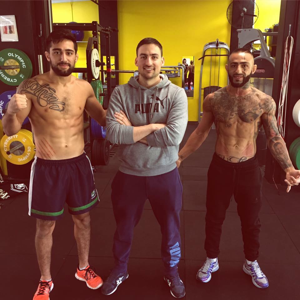 Three muscular men, including what appears to be a trainer, pose in a gym setting with weightlifting equipment in the background.