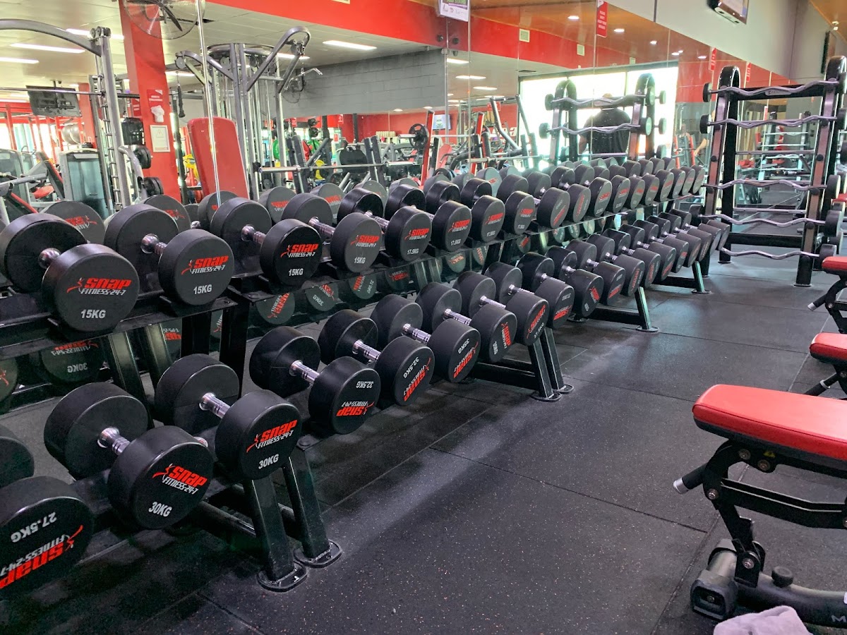 The photo showcases a row of dumbbell racks in a gym with red accents and mirrored walls.